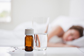 woman_sleeping_white_bed_with_bottle_of_sleeping_aids_pills_on_white_table_next_to_half_a_glass_of_water