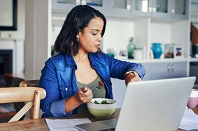 A young woman checking her watch while eating a salad and working on her computer from home.