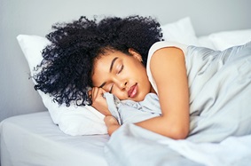 brown_skinned_young_woman_sleeping_in_bed_cuddled_with_grey_comforter