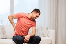 young adult male holds his left side while wincing in pain sitting on a white couch