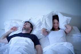 man snoring while in bed with a woman who is covering her head with a pillow
