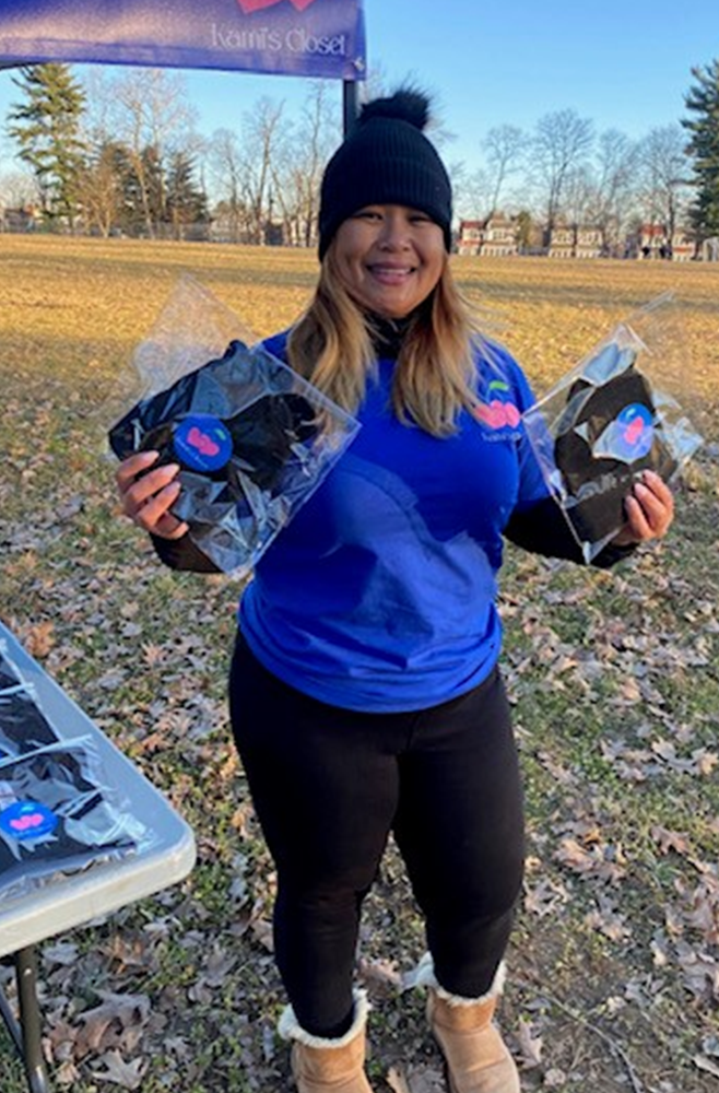 A woman stands outside under a blue tent, designed with the logo for Kami's Closet, holding a winter hat in each hand.